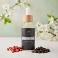 Bottle of 'The Anchor' dry body oil with a dropper on a light surface with flowers, pink peppercorn, and coffee beans in the background.